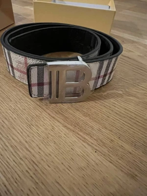 Burberry Bälte - Snyggt bälte från Burberry med klassiskt rutmönster i vitt, svart och rött. Bältet har ett stort silvrigt B-spänne framtill och är tillverkat i skinn. Perfekt accessoar för att lyfta din outfit.