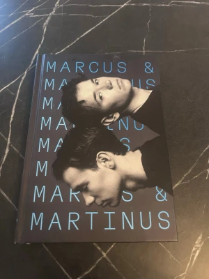 Marcus & Martinus bok - En bok med hårdpärm och svart omslag med blå text och ett foto på Marcus & Martinus. Perfekt för dig som gillar biografier om unga artister och vill veta mer om deras liv och karriär. Boken är tryckt på papper och har ett modernt, stilrent utseende.