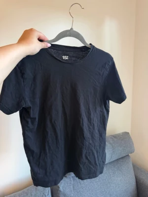 Svart basic t-shirt från 157 - En enkel svart t-shirt från 157 med rund hals och korta ärmar. Tillverkad i mjuk bomull och har en klassisk passform som funkar till allt. Perfekt basplagg för garderoben.