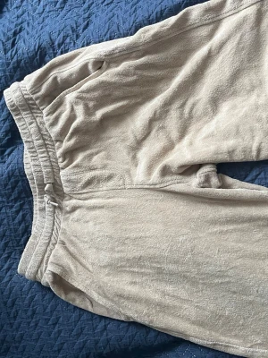 Beige mjukisbyxor med dragsko - Säljer ett par beige mjukisbyxor med resår i midjan och dragsko för skön passform. Byxorna har en bakficka och är gjorda i ett mjukt, frottéliknande material. Perfekta för chill dagar hemma eller när du vill vara extra bekväm.