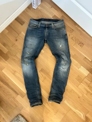 Blå slitna jeans från Nudie Jeans - Säljer ett par blå jeans från Nudie Jeans med slitningar och hål på benen för en cool, avslappnad look. Jeansen har klassisk femficksmodell, orangea sömmar och är tillverkade i denim. Perfekta för dig som gillar streetwear och en trendig stil. Modellen är tight long john. W27 L32