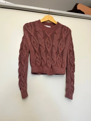 Rosa kabelstickad tröja från Zara - Trendig kabelstickad tröja från Zara i en dammig rosa nyans. Croppad modell med bred ribbad mudd i nederkant och vid ärmslut. Grovstickad struktur och rund halsringning. Perfekt för lager på lager och snygg till både jeans och kjol. Lite rost aktig färg 