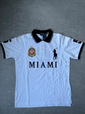 Polo Ralph Lauren Miami piké - Vit pikéskjorta från Polo Ralph Lauren med svart krage och detaljer. Stor broderad logga på bröstet, MIAMI-text och emblem i guld och rött. Kortärmad modell i bomull med knappar framtill och svarta detaljer på ärmarna.