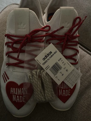 Adidas Human Made Tennis HU sneakers - Snygga vita Adidas Human Made Tennis HU sneakers med röda hjärtan och texten 'HUMAN MADE' på tån. Skorna har röda snören och extra vita snören medföljer. Ovandelen är i mesh för en luftig känsla och klassisk låg siluett. Aldrig använda. 