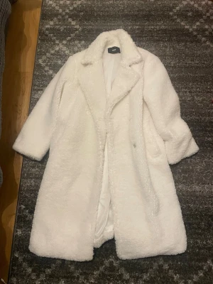 Vit lång teddyjacka med bälte - Supermysig vit teddyjacka från NA-KD med lång passform och bälte i midjan. Jackan har bred krage, dolda fickor och är gjord i fluffigt teddy-material som håller dig varm och stylish hela vintern.