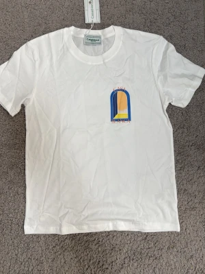 Vit Casablanca t-shirt med tryck - Säljer en vit t-shirt från Casablanca med färgglatt grafiskt tryck i blått, gult och orange på bröstet och ryggen. T-shirten har rund hals och korta ärmar, tillverkad i mjuk bomull. Perfekt för dig som gillar stilrena plagg med en twist.