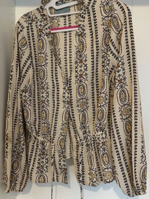 Mönstrad beige blus med knytband - Säljer en beige blus med mönster. Blusen har långa ärmar, knytband i midjan och vid halsen samt volangdetaljer nedtill. Materialet känns lätt och luftigt, perfekt för en avslappnad stil.