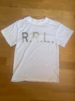 Vit RRL t-shirt med tryck - Säljer en vit t-shirt från RRL med stort grått tryck på bröstet. Klassisk passform med rund hals och korta ärmar. Perfekt till jeans eller shorts för en clean och enkel stil.