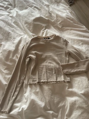 Beige långärmad topp med volangkant - Säljer en beige långärmad topp med volangkant vid hals, ärmslut och nederkant. Toppen har en enkel, slim passform och är tillverkad i mjuk bomull. Perfekt att styla med jeans eller kjol för en clean look.