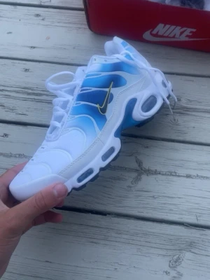 Nike Air Max TN - Nike Air Max Plus sneakers i vitt och blått med snygg gul Swoosh på sidan. Skorna har en gradient färgskala från vitt till blått, synlig Air Max-dämpning och detaljer i plast och mesh för extra komfort och stil. Perfekta för dig som vill sticka ut.