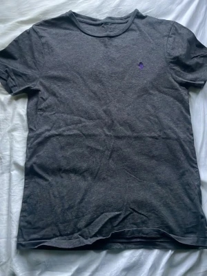 Mörkgrå t-shirt från Polo Ralph Lauren - Säljer en mörkgrå t-shirt från Polo Ralph Lauren med klassisk passform och korta ärmar. Liten lila logga broderad på bröstet. Perfekt basplagg till garderoben och funkar till det mesta.