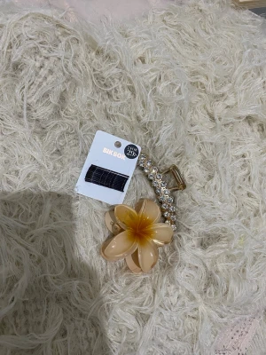Hårklämma med blomma och strass - Säljer en hårklämma från BIK BOK med guldfärgad metall, dekorerad med en stor beige och gul blomma i plast samt gnistrande strassdetaljer. Perfekt för att piffa upp frisyren med en söt och trendig accessoar.
