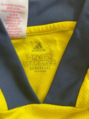 Svensk landslagströja Adidas gul/blå - Gul matchtröja från Adidas med blå detaljer och Sveriges landslagsemblem på bröstet. Tröjan har korta ärmar, blå krage och klassiska Adidas-ränder på axlarna. Tillverkad i lätt och ventilerande AEROREADY-material,storlek 152