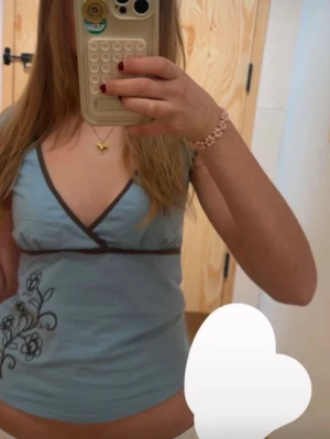 Ljusblå omlottopp med broderi från urban outfiter - Snygg ljusblå omlottopp med mörkbruna kanter och v-ringning. Toppen har ett broderat blommönster i svart på ena sidan och är ärmlös. Perfekt för dig som gillar en feminin och trendig stil. Fråga om du undrar något eller vill ha mer bilder!