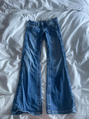 Blå bootcut jeans med fickdetaljer - Säljer ett par blå bootcut jeans med klassisk femficksdesign och snygga detaljer på bakfickorna. Jeansen har normalhög midja och utsvängda ben. Perfekta för dig som gillar retrovibbar och vill ha ett par jeans som sticker ut lite extra.