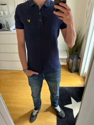 Marinblå piké från Lyle & Scott - Snygg marinblå pikétröja från Lyle & Scott med klassisk krage och knappslå. Tröjan har korta ärmar och den ikoniska gula logon broderad på bröstet. Tillverkad i mjuk bomull som känns skön mot huden. Perfekt till jeans eller shorts för en clean look.