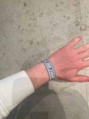 Blått tygarmband från Dior - Säljer ett blått tygarmband från Dior med texten 'J'ADIOR' invävt i vitt. Armbandet har justerbar knytning med tofsar i ändarna och en cool, avslappnad stil. Perfekt accessoar för att lyfta din outfit med en exklusiv touch.