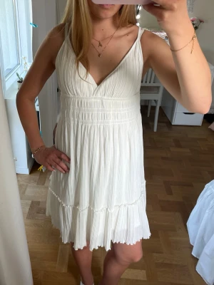 Vit kort sommarklänning med volang - Söt vit kortklänning med tunna axelband och v-ringning. Klänningen har plisserad kjol, volangdetalj nedtill och är tillverkad i ett lätt tyg som passar perfekt för varma dagar. Stilren och enkel design som ger en fräsch look.