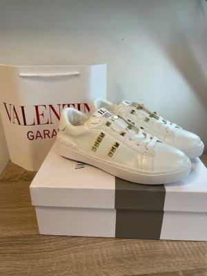 Valentino Rockstud vita sneakers - Snygga vita sneakers från Valentino med guldiga nitar längs sidorna och på plösen. Modellen heter Rockstud Untitled och har en klassisk låg siluett. Skorna är tillverkade i slätt läder och har vita skosnören samt en vit sula. Perfekta för dig som gillar exklusiv och trendig stil.