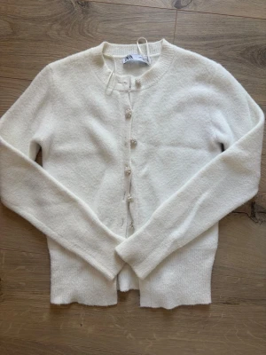 Vit kofta med pärlknappar från Zara M men liten  - Söt vit kofta från Zara med rund halsringning och dekorativa pärlknappar framtill. Koftan är stickad i mjukt material och har långa ärmar samt ribbade muddar vid ärmslut och nederkant. Perfekt för lager på lager och en clean look. Storlek M men passar S 