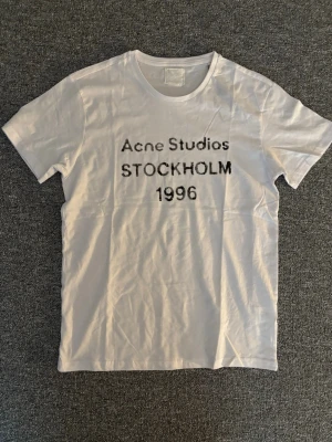 Vit Acne Studios t-shirt med tryck - Säljer en vit t-shirt från Acne Studios med svart texttryck 'Acne Studios STOCKHOLM 1996' på bröstet. Klassisk rund hals och korta ärmar. Tillverkad i mjuk bomull för en skön och avslappnad stil. ALLA lappar saknas därav priset