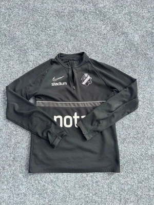 Svart AIK träningströja från Nike - Svart långärmad träningströja från Nike med AIK:s klubbmärke på bröstet. Tröjan har halv dragkedja, tryckta loggor från Stadium och Notar samt en tunn vit linje över bröstet. Perfekt för fotbollsträning eller sportiga aktiviteter. På tröjan står strlk 147-158cm men passar upp till 170cm