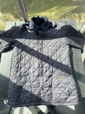 Quiltad jacka från Barbour, marinblå - Snygg marinblå quiltad jacka från Barbour med klassisk krage i manchester. Jackan har tryckknappar framtill och två stora fickor. Perfekt för dig som gillar stilren och tidlös design. 