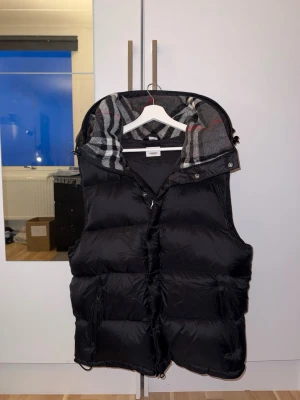 Burberry Väst/jacka - Säljer en stilren svart väst från Burberry med ikoniskt rutigt foder i kragen  Den kan enkelt göras om till en jacka – perfekt för både höst, vinter och vår.  * Avtagbara ärmar (2-i-1: väst + jacka) * Varm och lätt puffmodell * Klassisk Burberry-rutig insida i kragen * Dragkedja + knappar * Två fickor framtill