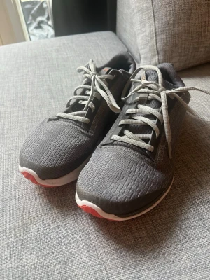 Gråa Under Armour sneakers - Snygga grå sneakers från Under Armour med mesh-ovandel och vita sulor. Skorna har gråa skosnören och en orange logga på hälen. Storlek 38. Tackvare sin ljusa sula så går det perfekt att använda skorna till inomhusträning. 