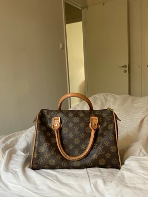 väska  - Säljer min vintage väska handväska i brunt monogramcanvas med ljusbruna läderdetaljer och guldfärgade metalldelar. Väskan har två handtag och dragkedja upptill. Rymmer mycket .  Perfekt för dig som gillar tidlös och lyxig design.