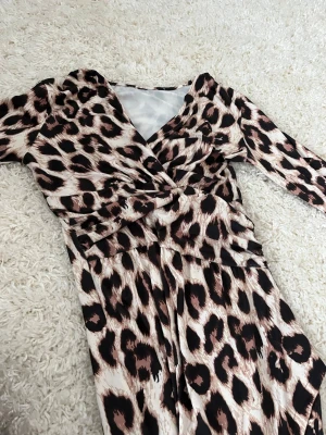 Leopardmönstrad långklänning med slits - Snygg långklänning med leopardmönster och v-ringning. Klänningen har långa ärmar och en markerad midja med draperad effekt framtill. Materialet känns mjukt och följsamt, perfekt för dig som vill sticka ut. Slits framtill för extra edge.