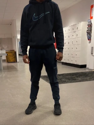 Mörkblå Nike mjukisbyxor med logga - Säljer ett par mörkblå mjukisbyxor från Nike med en stilren ljusblå swoosh-logga på benet. Byxorna har en slim fit-modell med muddar vid bensluten och dragsko i midjan. Perfekta för chill eller träning, tillverkade i mjukt och bekvämt material.