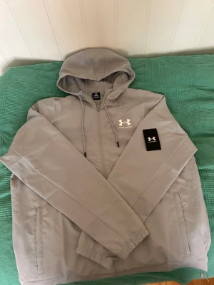 Grå vindjacka från Under Armour - Snygg grå vindjacka från Under Armour med huva och dragsko. Jackan har dragkedja framtill, två sidofickor och elastiska muddar vid ärmsluten. Tillverkad i lätt och vindtätt material, perfekt för blåsiga dagar. Under Armour-logga i vitt på bröstet.  Använd bara en gång helt nytt. 