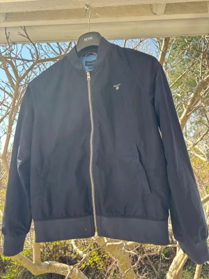 Mörkblå bomberjacka från Gant - Snygg mörkblå bomberjacka från Gant med dragkedja framtill och två fickor. Jackan har ribbade muddar vid ärmslut och midja samt diskret Gant-logga på bröstet. Perfekt för dig som gillar klassisk och stilren streetwear.