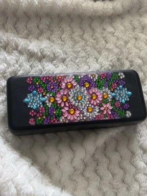 Svart glasögonfodral med blommor -  svart glasögonfodral dekorerat med färgglada blommor i rosa, lila, blått, grönt och silver diamanter. Fodralet är hårt och har en insida i ljusgrått tyg som skyddar dina solglasögon. 