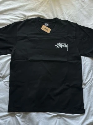 Svart Stüssy t-shirt med logga - Svart t-shirt från Stüssy med vit logga tryckt på bröstet. Klassisk rund halsringning och korta ärmar. Perfekt för en streetwear-look och enkel att matcha med andra plagg.