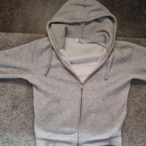 Grå zip hoodie från Bvaldi - Klassisk grå hoodie med dragkedja från Bvaldi. Hoodien har huva med dragsko, två fickor framtill och ribbade muddar vid ärmslut och nederkant. Perfekt för en avslappnad och enkel stil.