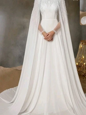 Vit långklänning med cape och pärldetaljer - Elegant vit långklänning med svepande cape och vackra pärldetaljer upptill. Klänningen har en klassisk, figurnära midja och ett långt, flödande fall. Perfekt för dig som vill ha en riktigt sagolik look.