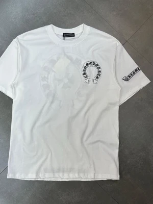 Vit t-shirt från Chrome Hearts - Vit t-shirt från Chrome Hearts med tryck på bröstet och ärmen. Klassisk rund halsringning och korta ärmar. Stilen är enkel men med tydliga detaljer från märket, perfekt för en streetwear-look.