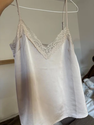 Champagnefärgad linnetopp med spets - Snygg champagnefärgad linnetopp med tunna axelband och v-ringning dekorerad med spets. Toppen har en silkeslen känsla och är perfekt för att skapa en chic och trendig look.