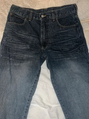 Levi's blå raka jeans - Klassiska blå jeans från Levi's med rak passform och fem fickor. Jeansen har slitningar och ljusare partier framtill för en cool, använd look. Tillverkade i bomull med Levi's ikoniska röda etikett på bakfickan.