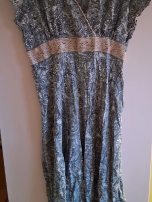 Blå paisleymönstrad klänning - Snygg blå klänning med paisleymönster och ljus spetsdetalj i midjan. Klänningen har korta ärmar och ett lätt, luftigt tyg som ger en avslappnad känsla. Perfekt för dig som gillar boho-stil och unika mönster.