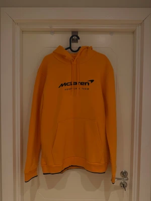 McLaren F1 hoodie - Snygg orange hoodie från McLaren Formula 1 Team med svart logga på bröstet. Hoodien har huva med dragsko, magficka och ribbade muddar. Perfekt för dig som gillar racing och vill sticka ut med en sportig stil.
