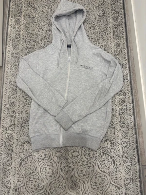 Ljusgrå zip hoodie från McKenzie - Ljusgrå hoodie från McKenzie med dragkedja framtill och justerbar huva med snören. Tillverkad i mjukt material och har ribbade muddar vid ärmar och nederkant. Diskret logga på bröstet för en clean look.