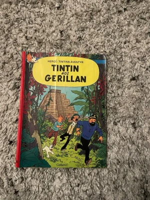 Tintin hos gerillan - Hergé - Säljer seriealbumet 'Tintin hos gerillan' av Hergé. Boken har färgglad hårdpärm med illustration av Tintin, Kapten Haddock och Milou i djungelmiljö framför en gammal ruin. Perfekt för dig som gillar klassiska äventyrsserier och samlar på Tintin.