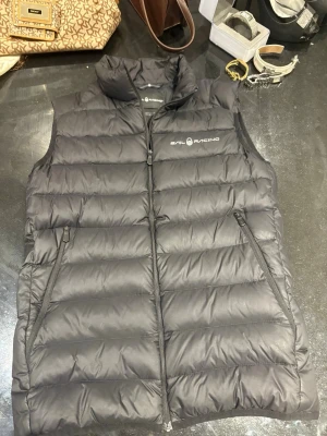 Svart dunväst från Sail Racing - Snygg svart dunväst från Sail Racing med hög krage och två dragkedjeförsedda fickor. Västen har en quiltad design och logga på bröstet. Perfekt för lager-på-lager och håller dig varm under kyliga dagar. Materialet är dun och polyester.