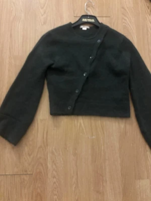 Mörkgrön asymmetrisk kofta - Snygg mörkgrön kofta med asymmetrisk knäppning framtill. Koftan har långa ärmar och en croppad passform som ger en modern look. Perfekt att styla över en topp eller klänning för extra lager. Mjuk och skön kvalitet.