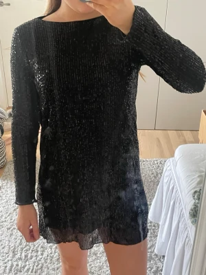 Svart glittrig kortklänning - Snygg svart kortklänning med långa ärmar och täckt av glittriga paljetter. Klänningen har en rak passform och rund halsringning. 