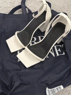 Vita chunky pumps med fyrkantig tå - Säljer ett par vita pumps från Rubi med chunky blockklack och fyrkantig tå. Skorna har en stilren design med ankelrem och är tillverkade i ett slätt syntetmaterial. Perfekta för dig som gillar trendiga och bekväma klackar.