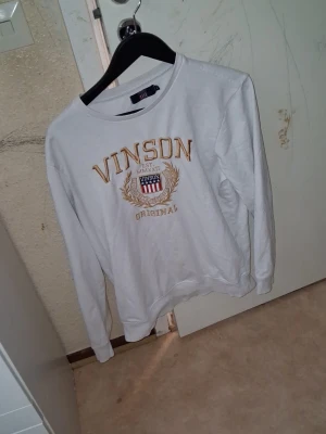 Vit sweatshirt från Vinson (Äkta) - Vit sweatshirt från Vinson med guldfärgad brodyr och tryck framtill. Rund halsringning och långa ärmar. Klassisk college-stil med mjukt material och ribbade muddar vid ärmslut och nederkant. Perfekt för en avslappnad look.